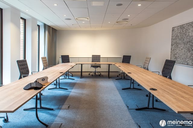 Moderne vergaderruimte met U-vormige houten vergadertafel, grijze bureaustoel en blauwe vloerbedekking.