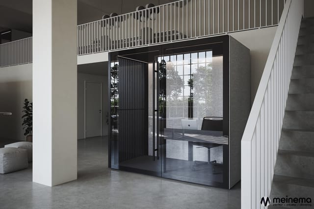 Moderne belcel met zwart frame en glazen wanden in kantoorruimte met betonnen vloer, witte zuilen en metalen trap.