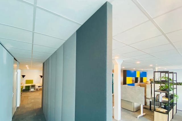 Modern kantoor met grijze akoestische scheidingswand, witte plafondpanelen, houten vloer, zithoeken met gele accenten en blau