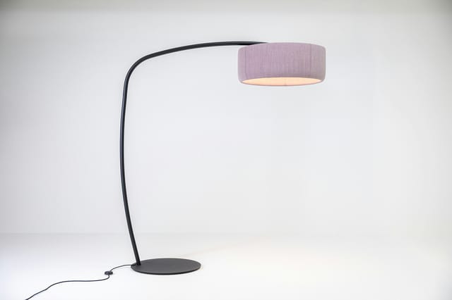 BuzziJet vloerlamp met een zwart gebogen frame, ronde voet en lichtroze cilindervormige kap tegen een witte achtergrond.