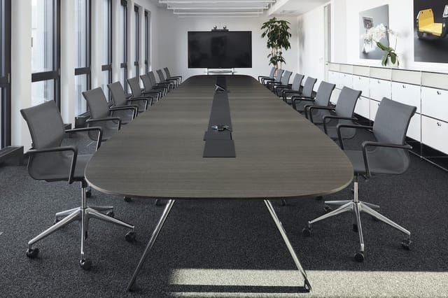 Moderne vergaderruimte met ovale grijze tafel, zwarte stoelen met wielen, grote wandscherm, glazen wanden en witte kasten.