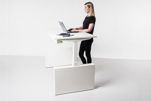 Vrouw werkend aan wit verstelbaar bureau met laptop in staande positie, geplaatst op wit onderstel in minimalistisch interieu