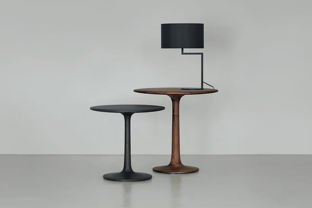 Twee ronde pedestal-tafels: één zwart metaal, één houtkleurig, met daarop een zwarte tafellampe met rechthoekige kap.