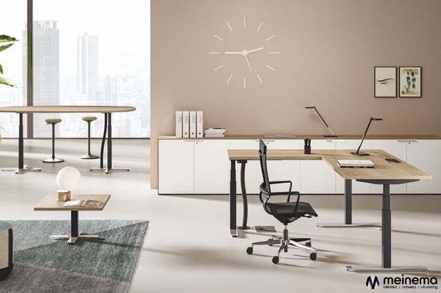 Modern kantoor met houten bureau's, zwarte bureausstoel, witte kasten, ronde tafel met barkrukken, grijze bank, wandklok en g
