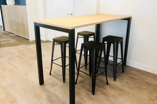 Bartafel met licht houten blad en zwart stalen frame, omgeven door vier bijpassende zwarte barkrukken in een kantoorruimte.