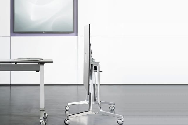 Mobiele displaystandaard op wielen in wit metaal met zwart scherm, in modern kantoorinterieur met whiteboard en tafel.