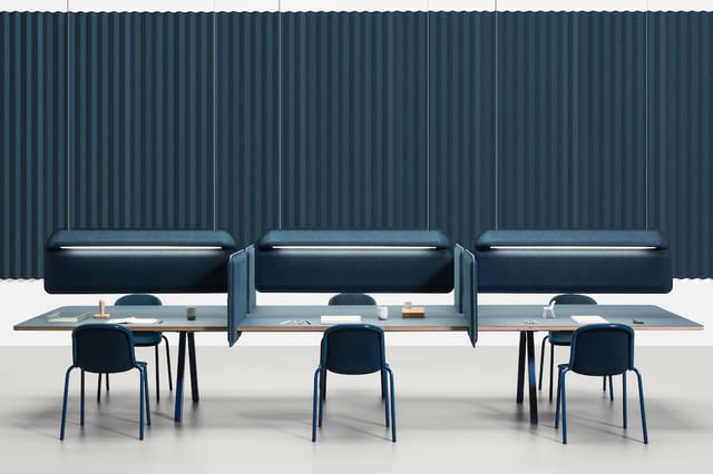 Moderne kantoorinrichting met drie donkerblauwe boxen met verlichte bovenkant tegen geribbelde muur, daarvoor drie werkplekke