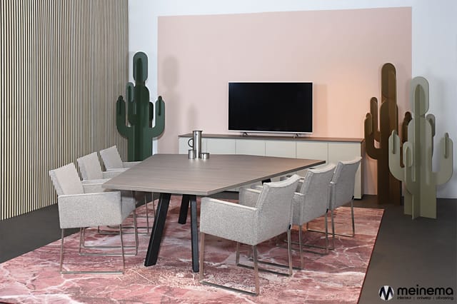 Vergaderkamer met grijze stoelen rond houten tafel, tv-meubel met scherm, cactusbeelden, roze muur en gestreepte wand.