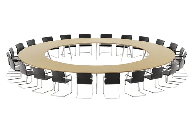 Grote ronde vergadertafel in licht hout met chromen onderstel, omringd door zwarte stoelen met metalen frame.