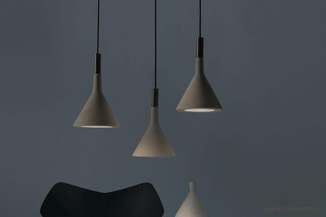 Drie betonnen hangende lampen in grijs/taupe met zwarte ophanging, tegen grijze wand, met zitmeubel onderaan.