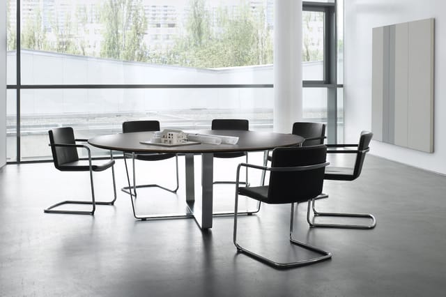 Ronde zwarte vergadertafel met zes zwarte kantooorstoelen met metalen frames in moderne kantoorruimte met grote ramen.