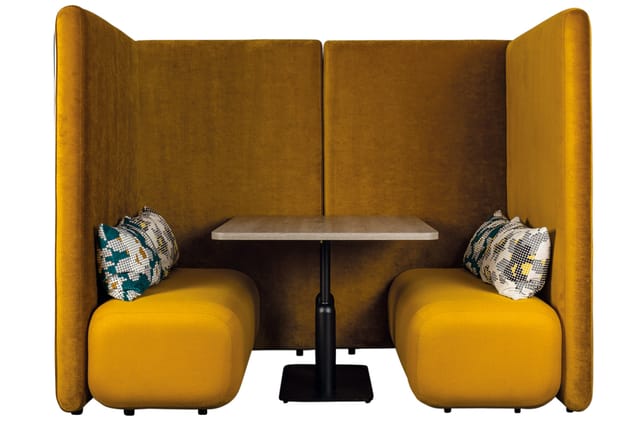 Gele privacy booth met twee zitplaatsen, gevoerde wanden, tafel tussen stoelen, decoratieve kussens.