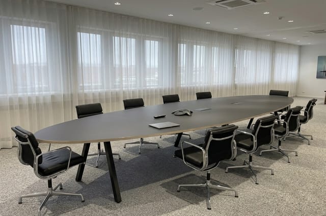 Moderne vergaderzaal met grijze ovale tafel, zwarte design stoelen, raampartij met wit gordijn en recessed lighting.