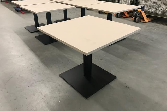 Vergadertafel met crèmekleurig blad en zwart onderstel in industriële ruimte met heftrucks op achtergrond.