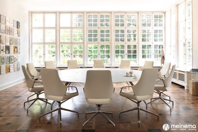 Ronde vergadertafel met beige conferentiestoel in moderne kantoorruimte met grote raampartijen en houtparketvloer.