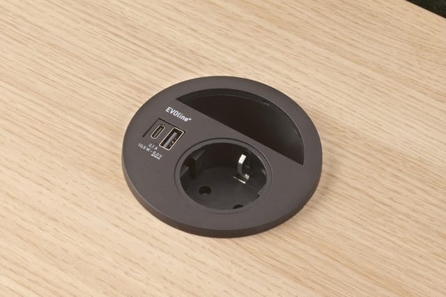 Zwart rond bureaustopcontact ingebouwd in lichte houten tafelblad met twee USB-poorten en één stopcontact.