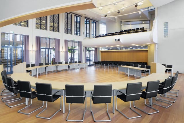 Grote vergaderruimte met ronde houten tafel, zwarte kantelstoelen, houten vloer, grote ramen, gele en witte wanden, tweede ni