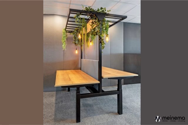 Houten vergadertafel onder zwart stalen pergola met hangende planten en verlichte pendels in kantooromgeving met grijze akoes