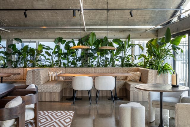 Moderne lounge met beige hoekbank, witte stoelen, ronde tafels en groene kantoorplanten tegen betonnen plafond.