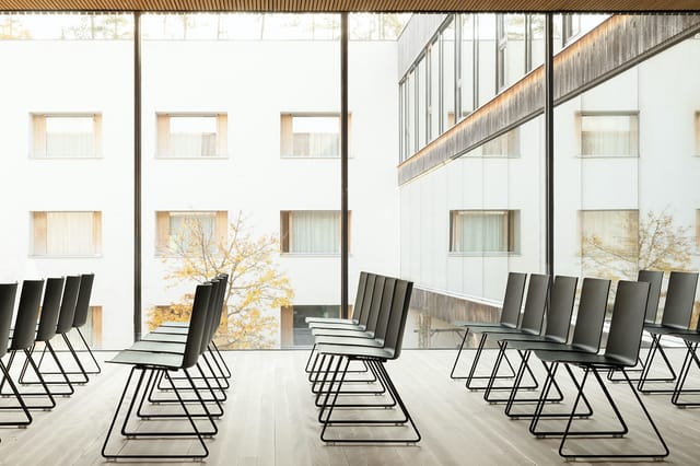 Moderne vergaderruimte met zwarte kantelstoelen op metalen frames, grote ramen, witte wanden en houten accenten.