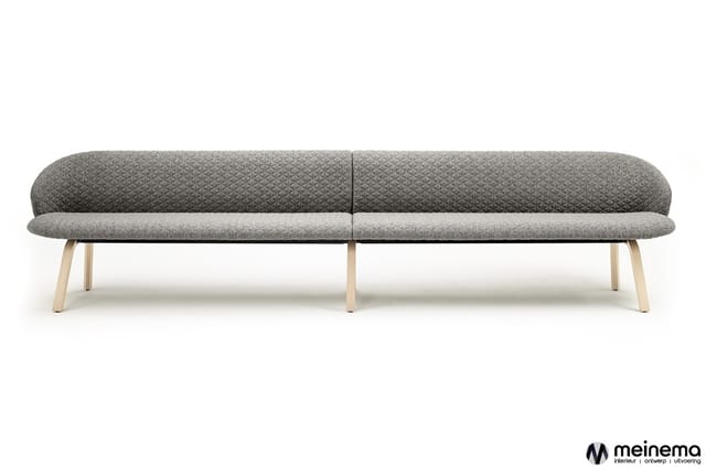 Grijze loungbank met afgerond design, gestoffeerd in gewatteerd textiel, op lichte houten poten. Meinema-logo onderin rechts.