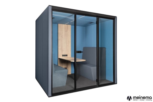 Gesloten belcel met zwart frame, blauwe wanden, houten bureau aan linkerzijde en grijze zitbank rechts.
