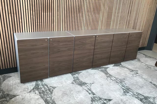 Modulaire houten kasten met witte bovenbladding, zes deuren in donker houtfineer, opgesteld tegen verticale wandpanelen in ka