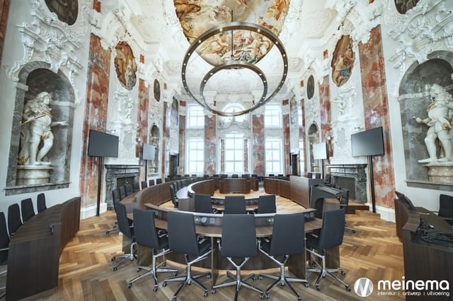 Luxe barokvergaderruimte met ronde houten vergadertafel, zwarte ergonomische bureaustoelen, witte stucplafond, rode wandschil