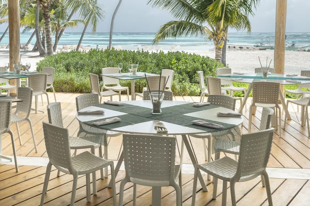 Grijze kunststof stoelen rond witte vergadertafel op houten terras met zee en palmen op achtergrond.