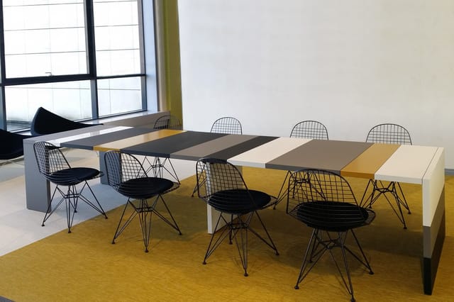 Vergadertafel met kleurvakken grijs/beige/geel en zwarte draaistoelen op geel tapijt in kantoorruimte met ramen.