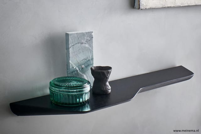 Zwarte wandplank met groene glazen doos, grijs-blauwe decoratieve blok en zwart object erop.