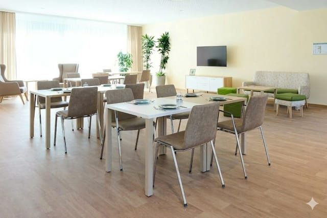 Modern kantoorruimte met twee rechthoekige tafels, grijze stoelen, houten vloer, beige wanden, groen accent, planten en TV.
