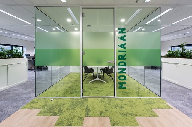 Glazen belcel met groene frosting en "HYDRAAN" tekst, twee zwarte stoelen, groen tapijt, houtlook vloer in modern kantoor.