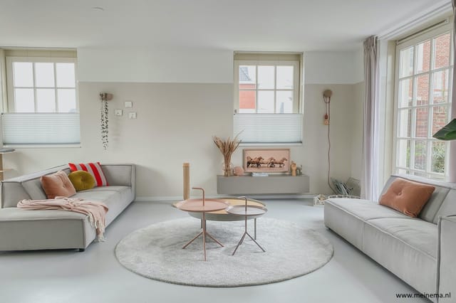 Moderne woonkamer met twee grijze banken, rond wit vloerkleed, twee ronde tafels in roze/wit, wandmeubel met framed artwork,