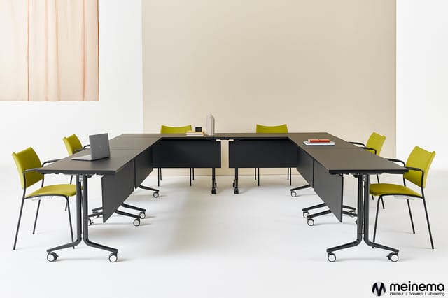 U-vormige vergadertafel in grijs met wielen, omgeven door gele stoelen met zwart frame en wielen, tegen beige wand.