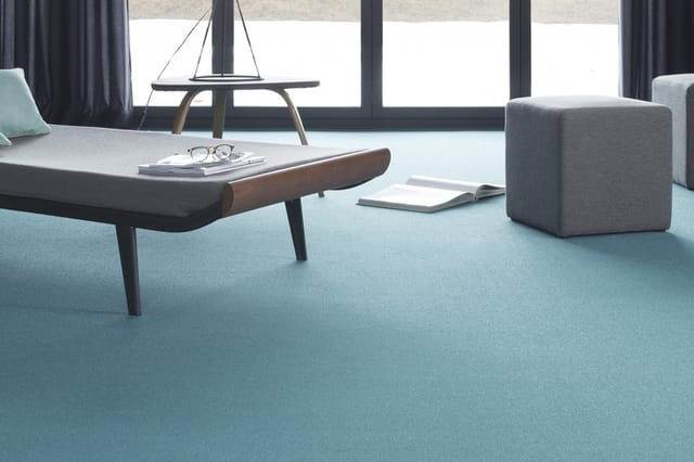 Licht kantoor met turquoise vloerbedekking, grijze bank, laag tafel met houten armsteun, grijze poef en grote ramen met grijz