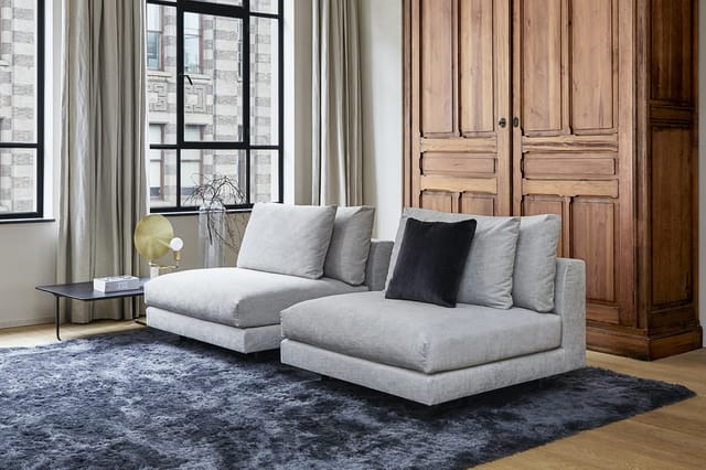 Grijze modulaire loungeset met chaise longue, kussens en zwart accent kussen in kantoor/woonruimte met houten kasten en raam.