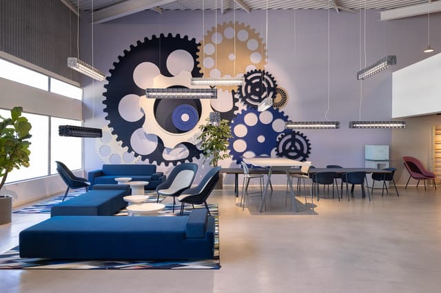 Modern kantoorinterieur met blauwe hoekbank, grijze stoelen, vergadertafel en grote muursculptuur met zwarte en gouden tandwi