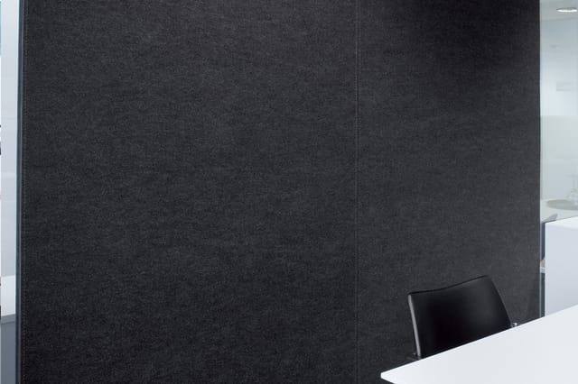 Donkergrijs akoestische wandpaneel in kantooromgeving met witte tafel en zwarte stoel op voorgrond.
