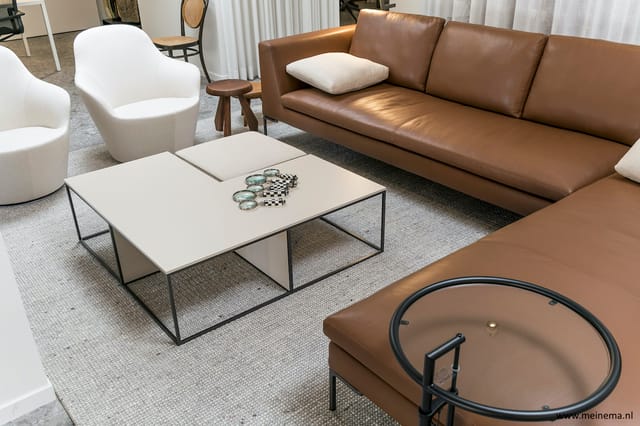 Showroom met bruine leren sofa, witte fauteuils, witte salontafel met metalen frame en decoratieve items, grijze tapijt.