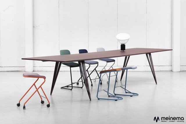 Vergadertafel met donker blad en zwart onderstel, omgeven door diverse mobile stoelen in rood, zwart, blauw en grijs. Witte i