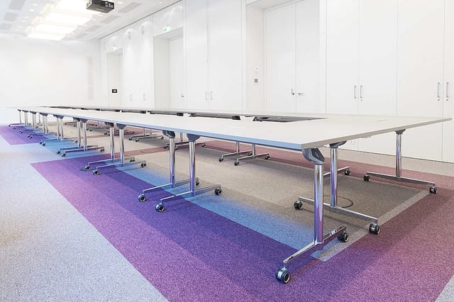Moderne vergaderruimte met vier witte rechthoekige tafels op wielen, zilveren metalen onderstel, paarse en blauwe tapijttegel